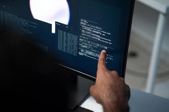 A software developer points at programming code displayed on a computer screen, reviewing data logs and source code while working on a technology project in an office.