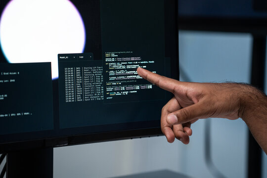 A person points at computer code displayed on a monitor while reviewing machine learning logs and technical data during a software development task in a modern office workspace.