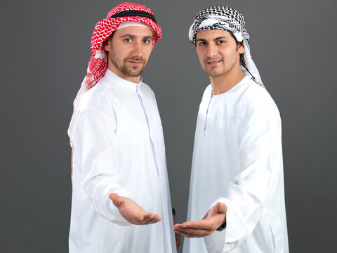 Two Arab men in traditional white kandura offering and holding empty hands open gesture isolated on grey background.