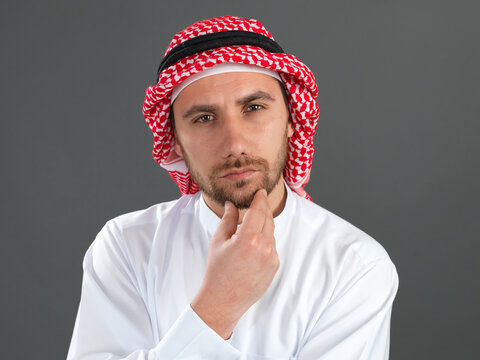 Close up portrait of thoughtful Arab man in traditional white kandura holding hand on chin isolated on grey background.