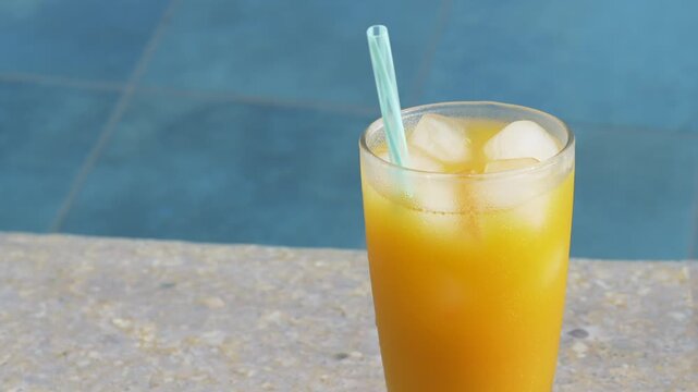 Chilled citrus cocktail waits in glass near pool edge, glowing orange with ice and straw. Refreshing drink presentation gives nonalcoholic cocktail bright summer feel with citrus juice and ice cubes.