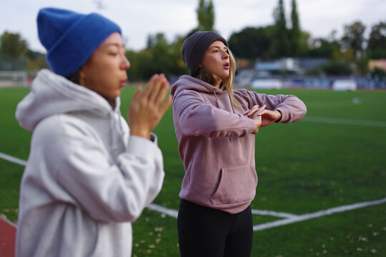 Two diverse young women are stretching their arms and bodies, warming up before exercising on a stadium track, representing friendship, teamwork, and an active lifestyle