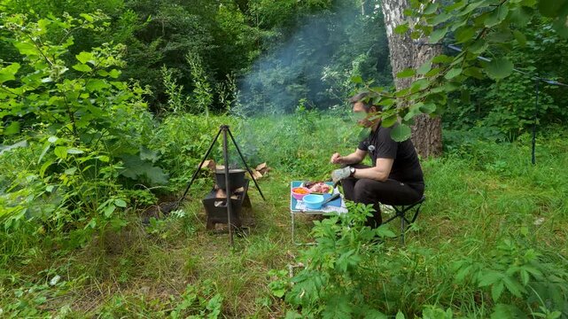 Man cooking food in cauldron over campfire during outdoor forest camping picnic lifestyle