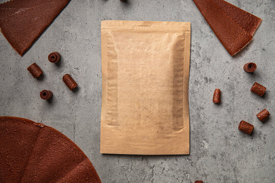 Natural fruit leather rolls and sheets arranged around craft paper ziplock pouch mock up on grey concrete background