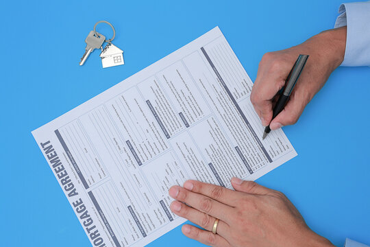 Focused hand signs mortgage agreement document ensuring loan approval and property ownership with keys nearby on blue background