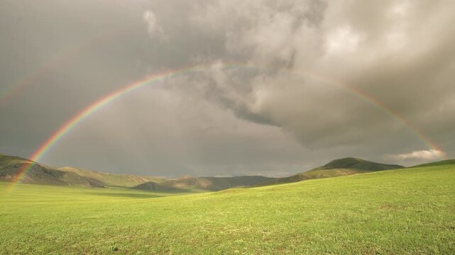 Rainbow arches above green hills and meadow after rain beneath breaking storm clouds.