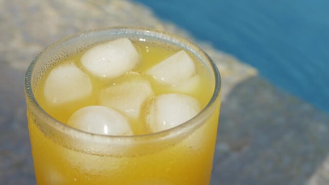 Refreshing orange beverage with ice cubes stands beside blue swimming pool, evoking carefree vacation leisure. Bright summer drink creates relaxed hospitality imagery for resort advertising and menus.