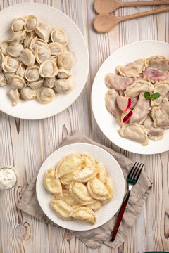 Three white plates with assorted cooked vareniki with different fillings and meat dumplings on rustic wooden background top view