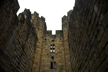 Looking up at high medieval stone castle walls and towers against white sky. Historic fortress architecture, ancient defensive structure. Alnwick castle England © TomKorcak