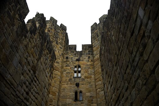 Looking up at high medieval stone castle walls and towers against white sky. Historic fortress architecture, ancient defensive structure. Alnwick castle England