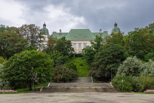 This scenic view captures the historic Ujazdow Castle nestled within lush green trees above a peaceful pond in Warsaw, Poland, showcasing the city's beautiful architectural heritage and nature