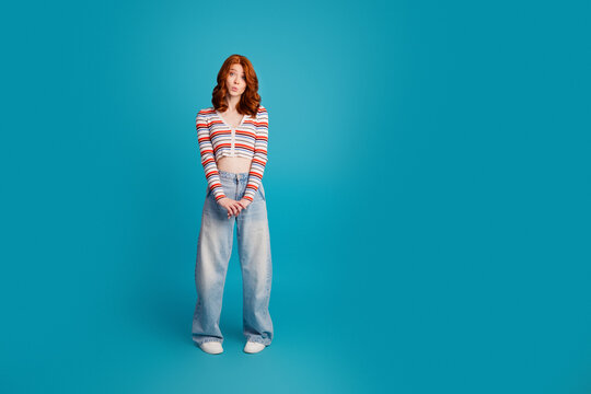 Young redhead woman standing in casual striped crop top and wide-leg jeans on turquoise background, playful surprised expression, fashion portrait and lifestyle concept