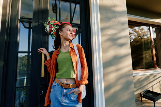 Young woman standing outdoors near a building entrance, smiling and looking away. She wears a vibrant orange shirt and blue jeans. Lifestyle concept for urban fashion and leisure.