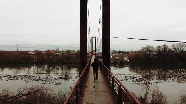 An old, rusty bridge over a murky river against an autumn backdrop, with a person walking across it.