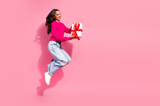 Young woman in pink sweater jumps with gift box against pink background for festive promotion and holiday shopping