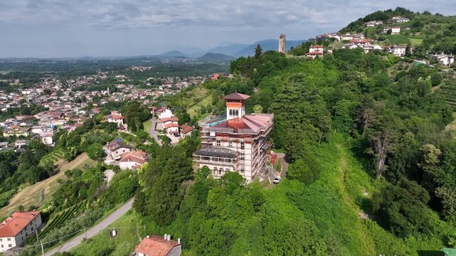 Drone overlooks historic stone ruins of Castello di Coia on green hill near Tarcento. Ancient tower remnant stands surrounded by dense forests and mountain backdrop in Italian countryside.