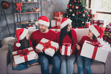 Naklejka premium Family celebrating Christmas at home exchanging gifts on sofa with Santa hats and decorated tree in cozy living room