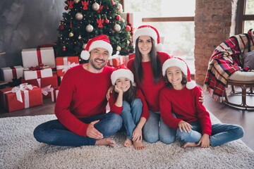 Naklejka premium Happy family in matching red sweaters and Santa hats sitting by a decorated Christmas tree with wrapped gifts, smiling together in a cozy holiday living room