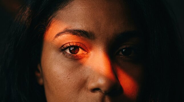 Extreme close-up of a woman&rsquo;s eyes crossed by a diagonal beam of saturated orange-red light, deep black shadows, cinematic beauty portrait photography, warm copper skin, sharp catchlights, graphic
