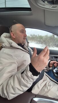 Middle aged man driving luxury vehicle through snowy forest landscape while gesturing during conversation. Fixed medium shot from the passenger seat.