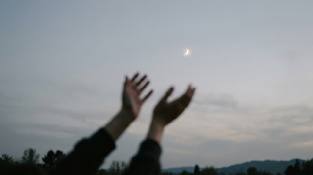 Two intentionally blurred hands reaching toward a tiny crescent moon in a pale evening sky, conceptual photography, shallow focus on the celestial point, soft gray-blue palette, minimalist