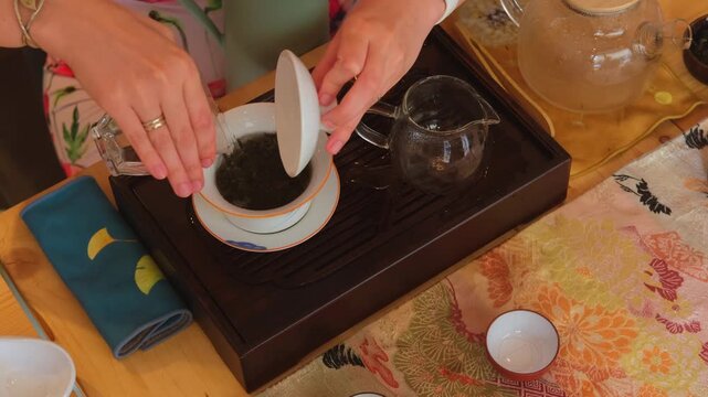 Tea preparation scene with leaves being added to a teapot on a wooden tray, water poured for brewing, warm daylight, calm ritual, domestic tea making, mindful moment, kitchen table setting