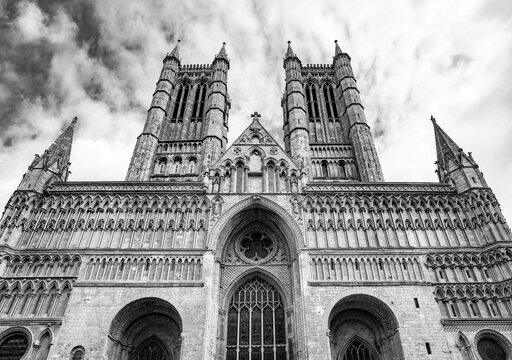 Monumental west front of Lincoln Cathedral in Lincoln, Lincolnshire, England, photographed from a low angle in high-contrast black and white beneath a dramatic cloudy sky.