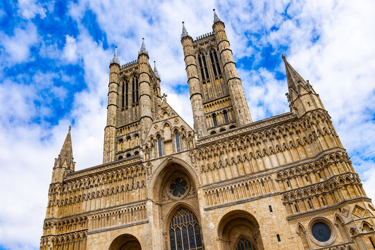 Low-angle wide-angle view of the monumental west front and towers of Lincoln Cathedral in Lincoln, Lincolnshire, England, beneath a vivid blue and cloudy summer sky.
