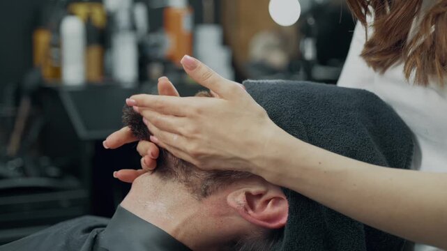 Barber applies lotion to a male clients beard during a professional grooming treatment in a barbershop.