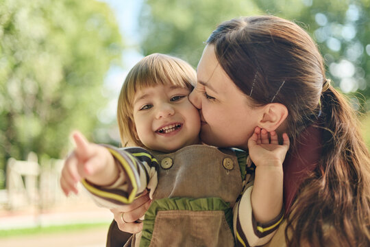 Caucasian young adult mother kissing cheerful toddler daughter outdoors in park. Imagery suitable for family lifestyle advertising, parenting campaigns, child care promotions