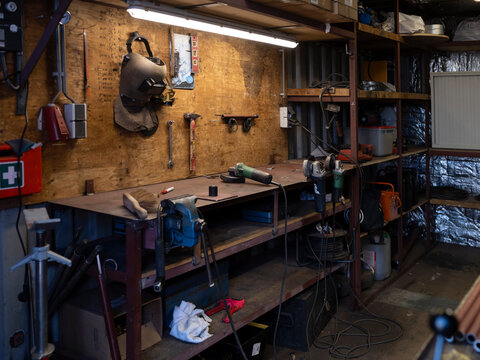 A steel workbench with vice, angle grinders and a welding helmet fills a container workshop lit by a single fluorescent tube.