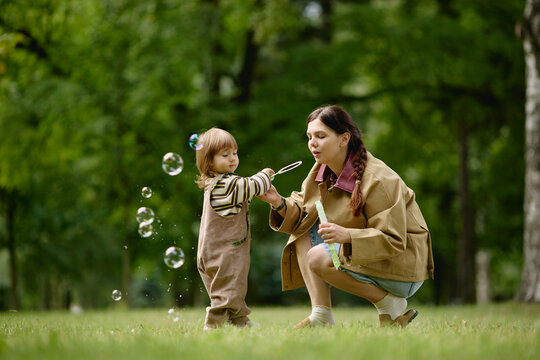 Caucasian young adult mother blowing soap bubbles with toddler child in park representing parenting and childcare. Visual suited for family lifestyle campaigns, nursery or preschool ads