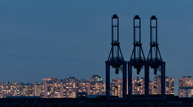 Industrial cargo cranes silhouette standing tall against a glowing residential apartment skyline during deep blue twilight
