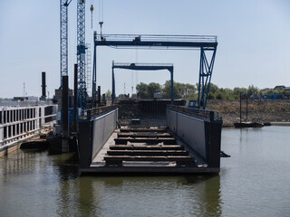 A blue gantry crane spans a dry dock at a river shipyard, a white river cruise ship lying alongside. © Victor