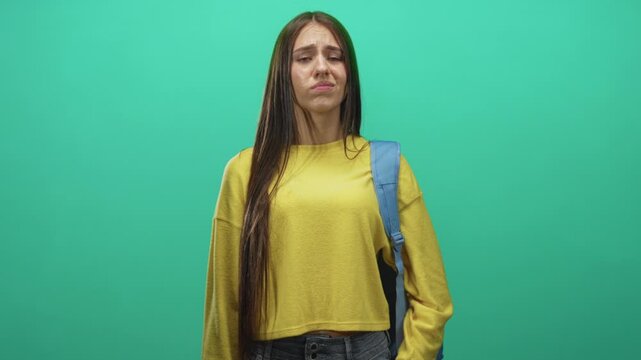 Young woman student wearing a yellow sweater and backpack grimaces with a raised hand gesture against a green studio wall; disgust.