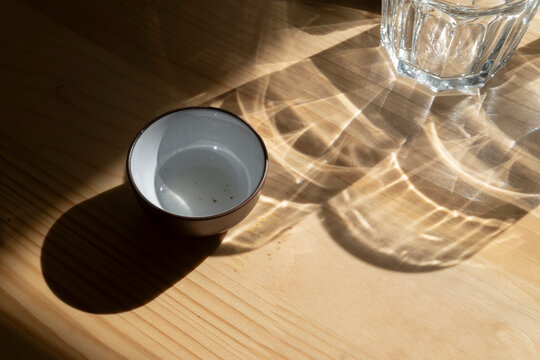 Cup on wood table with glass of water casting shadow and light pattern, minimal still life focusing on everyday objects and natural illumination in calm composition