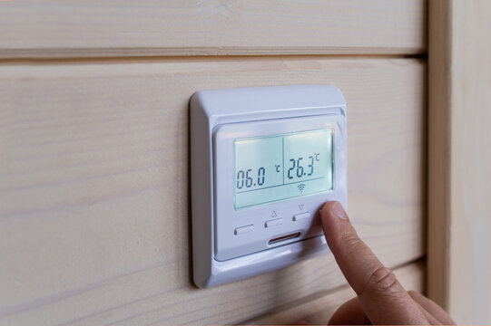 Woman adjusts wall-mounted digital thermostat by pressing control button to regulate indoor temperature and maintain comfortable home climate