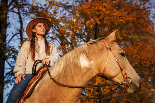 Cowgirl horseback riding in autumn nature during sunset. Woman in cowboy hat sitting on horse enjoying western lifestyle
