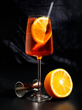 Classic Aperol Spritz cocktail in wine glass with ice cubes and orange slice, served with halved orange and bartending jigger on dark background. Perfect for summer aperitif and Italian drink concepts