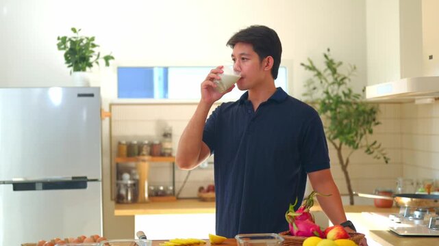 Young Asian Man Holding Glass of Milk in Modern Home Kitchen with Fresh Fruits on Counter