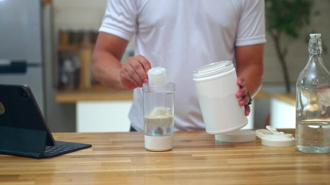 Closeup Fit man preparing protein shake with electric blender cup and protein powder container on wooden kitchen counter