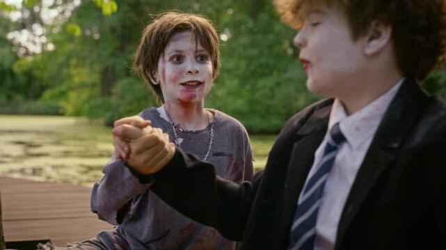 Caucasian teenagers wearing zombie makeup performing secret handshake outdoors by lake. Perfect for Halloween holiday marketing, youth event promotions, and spooky festive campaigns