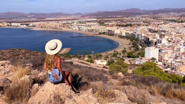 tourist woman with sun hat sitting on hill rock overlooking coastal town bay and mountains on sunny day- Aguilas in Spain, Murcia