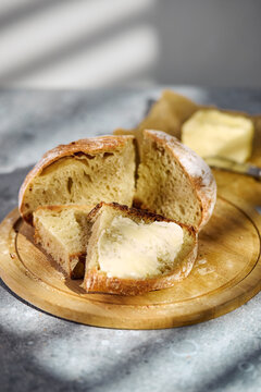 Close-up of freshly baked artisan sourdough bread sliced on a round wooden board. A slice topped with butter in the foreground, rustic food styling.