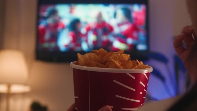 Young woman relaxing on a sofa at home in the evening, watching movies or TV series after work. Holding a bucket of chips and enjoying entertainment and leisure time. High quality 4k footage