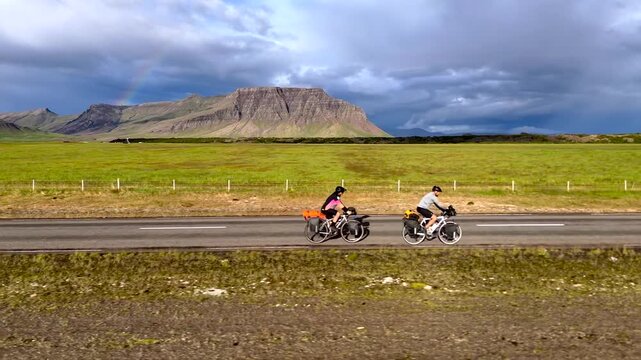 Two Bikepackers Cycling Along Iceland Highway Under Rainbow and Mountain Skyline