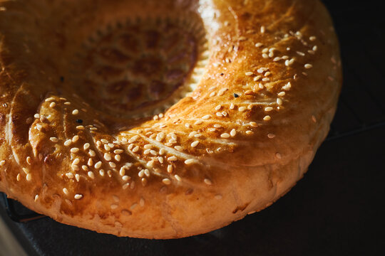 Close-up of golden Uzbek non bread with decorative center pattern and sesame seeds. Traditional Central Asian flatbread on dark background, fresh homemade baking.