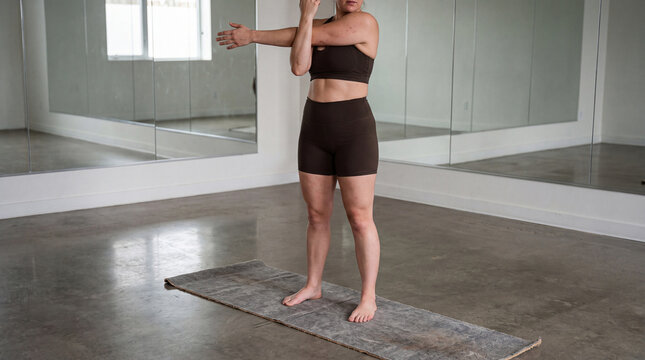 Woman performing a shoulder stretch on a yoga mat in a bright fitness studio with mirrors, focusing on flexibility, warm up, and mindful movement for a healthy routine