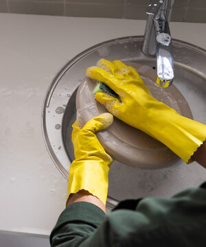 Gloved hands are scrubbing beige lid in stainless sink with green sponge, soap suds, chrome faucet