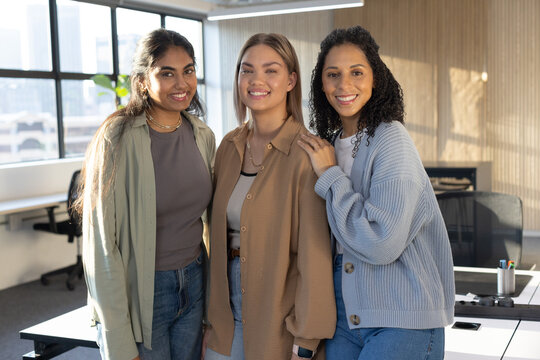 Diverse female coworkers standing smiling in office wearing cardigans and jeans with pen cup